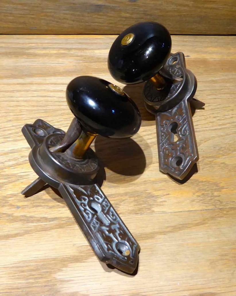 Closeup of doorknobs - Antique Eastlake passage set with gravity lock. Features porcelain doorknobs built into bronze plated patterned backplates, and brass patterned faceplate, set comes with strike plate.